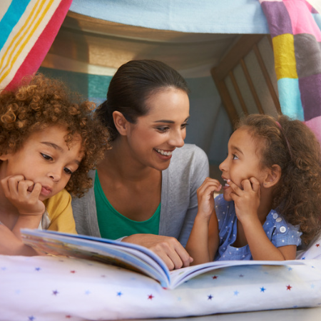 Image of mom and two children reading a book in a blanket fort.
